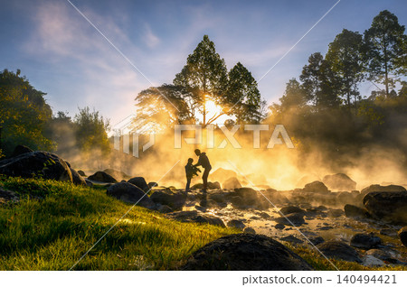 Beautiful sunrise at a misty hot spring surrounded by rocks and trees. A mother and child enjoy the golden light and warm, tranquil atmosphere in this serene natural setting. 140494421