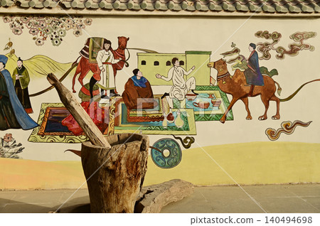 An ancient mortar and pestle by the wall, adorned with paintings depicting life in the Kuche Palace, China. An ancient mortar and pestle by the wall, adorned with paintings depicting life in the Kuche Palace, China. 140494698