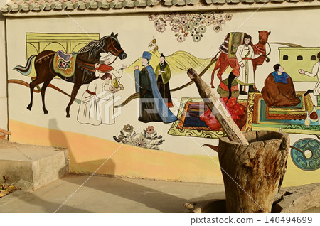 An ancient mortar and pestle by the wall, adorned with paintings depicting life in the Kuche Palace, China. 140494699