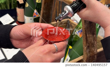畫面中,人們將清酒倒入紅色的杯子中,舉杯祝酒,讓人聯想到日本的飲酒聚會和飲食文化。 畫面中,人們將清酒倒入紅色的杯子中,舉杯祝酒,讓人聯想到日本的飲酒聚會和飲食文化。 140495157