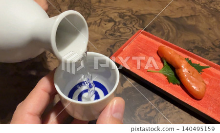 A scene of enjoying a drink in the evening, pouring chilled sake from a sake cup; Japanese food and sake enjoyed with seared cod roe. A scene of enjoying a drink in the evening, pouring chilled sake from a sake cup; Japanese food and sake enjoyed with seared cod roe. 140495159