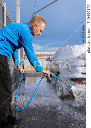 Man Washing Car at Self Service Car Wash. Applying thick foam with a pressure washer. The scene takes place in an outdoor washing bay with modern self-service equipment, automotive accessories Man Washing Car at Self Service Car Wash. Applying thick foam with a pressure washer. The scene takes place in an outdoor washing bay with modern self-service equipment, automotive accessories 140499193