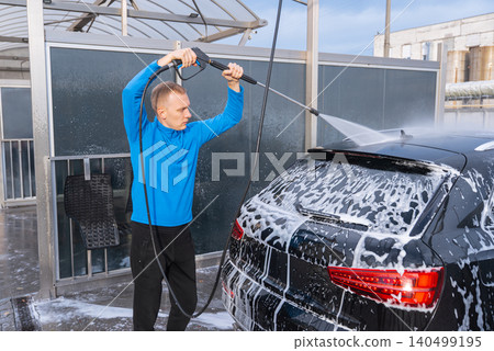 Man cleans his vehicle at self service car wash station using professional washing equipment. Rinsing the vehicle with high-pressure water stream system. Outdoor washing bay Man cleans his vehicle at self service car wash station using professional washing equipment. Rinsing the vehicle with high-pressure water stream system. Outdoor washing bay 140499195
