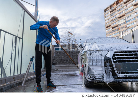 Man Washing Car at Self Service Car Wash. Applying thick foam with a pressure washer. The scene takes place in an outdoor washing bay with modern self-service equipment, automotive accessories 140499197