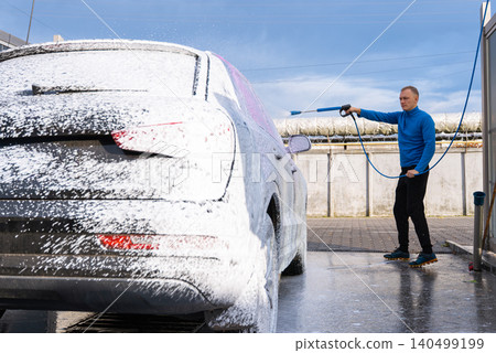 Man Washing Car at Self Service Car Wash. Applying thick foam with a pressure washer. The scene takes place in an outdoor washing bay with modern self-service equipment, automotive accessories Man Washing Car at Self Service Car Wash. Applying thick foam with a pressure washer. The scene takes place in an outdoor washing bay with modern self-service equipment, automotive accessories 140499199