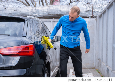 Man cleans his vehicle at self service car wash station drying and wiping the exterior with microfiber rags. Outdoor washing bay with modern self-service equipment, automotive accessories Man cleans his vehicle at self service car wash station drying and wiping the exterior with microfiber rags. Outdoor washing bay with modern self-service equipment, automotive accessories 140499211