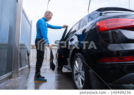 Man cleans his vehicle at self service car wash station cleaning removable car mats. Outdoor washing bay with modern self-service equipment, automotive accessories and cleaning tools. Modern Man cleans his vehicle at self service car wash station cleaning removable car mats. Outdoor washing bay with modern self-service equipment, automotive accessories and cleaning tools. Modern 140499236