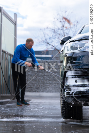 Man cleans his vehicle at self service car wash station using professional washing equipment. Rinsing the vehicle with high-pressure water stream system. Outdoor washing bay Man cleans his vehicle at self service car wash station using professional washing equipment. Rinsing the vehicle with high-pressure water stream system. Outdoor washing bay 140499249