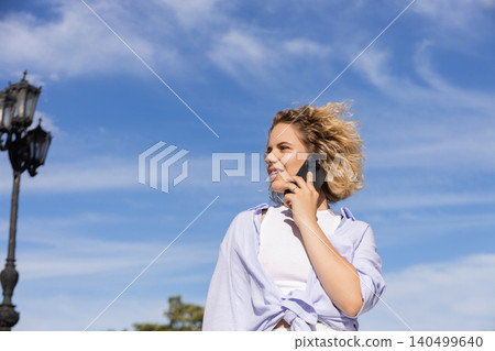 Woman smiles while talking on phone outdoors with blue sky Woman smiles while talking on phone outdoors with blue sky 140499640