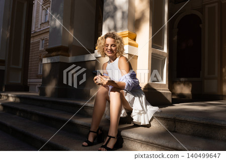 Person sitting on steps, smiling while using smartphone Person sitting on steps, smiling while using smartphone 140499647