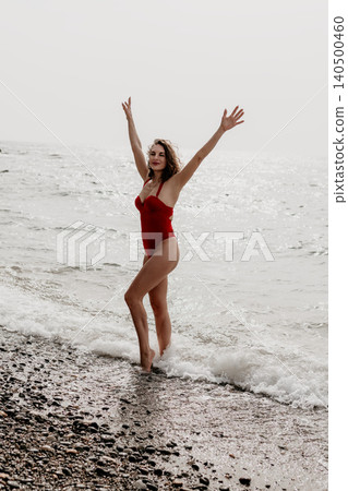 Woman beach swimsuit stands on rocky shore with arms raised as waves crash around her feet enjoying summer vacation 140500460