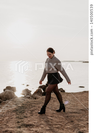 Woman walking coast young stylish woman in gray sweater and black skirt walks along a rocky shoreline near calm water Woman walking coast young stylish woman in gray sweater and black skirt walks along a rocky shoreline near calm water 140500539