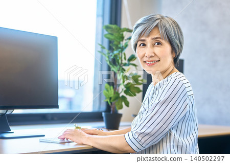 A gray-haired woman smiles as she types on her keyboard at a desk by the window. 140502297