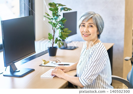 A gray-haired woman smiles as she types at a desk by the window. A gray-haired woman smiles as she types at a desk by the window. 140502300