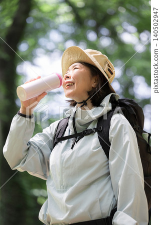 An active woman with a smile enjoying trekking; drinking water to stay hydrated; an image of an outdoor scene in early summer. An active woman with a smile enjoying trekking; drinking water to stay hydrated; an image of an outdoor scene in early summer. 140502947