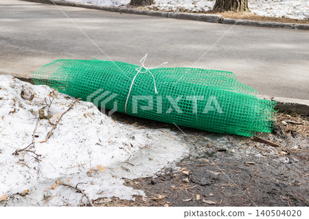 A Thick Roll of Green Plastic Netting Material Prepared for Landscape Protection and Project Use Next to a Curb on a Street Amidst Snow and Leaves A Thick Roll of Green Plastic Netting Material Prepared for Landscape Protection and Project Use Next to a Curb on a Street Amidst Snow and Leaves 140504020