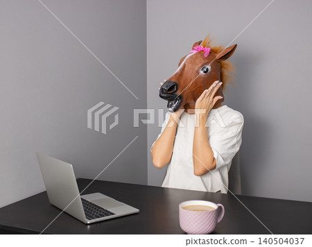 Office worker wearing horse head mask using laptop at desk with coffee cup Office worker wearing horse head mask using laptop at desk with coffee cup 140504037