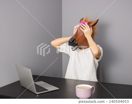 Office worker wearing horse head mask while sitting at desk with laptop and coffee mug in home office 140504053