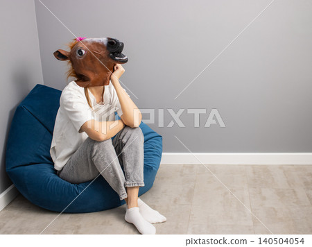 Adult wearing horse head mask sitting on blue bean bag chair in minimalist room with copy space Adult wearing horse head mask sitting on blue bean bag chair in minimalist room with copy space 140504054