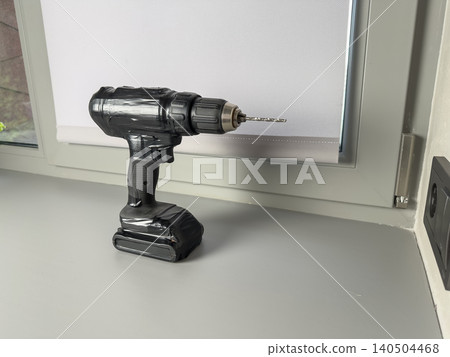 A Close-Up View of a Modern Cordless Drill Positioned on a Clean Work Surface Near a Window, Highlighting Its Functional Design and Quality Craftsmanship A Close-Up View of a Modern Cordless Drill Positioned on a Clean Work Surface Near a Window, Highlighting Its Functional Design and Quality Craftsmanship 140504468