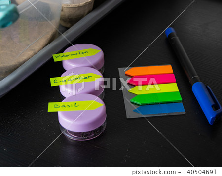 Labeled seed jars for watermelon, cucumber and basil on desk with pen and color page markers Labeled seed jars for watermelon, cucumber and basil on desk with pen and color page markers 140504691