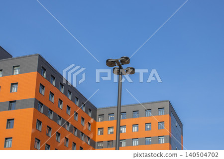 Modern Urban Building with Bright Orange Facade and Street Lighting Against Clear Blue Sky Modern Urban Building with Bright Orange Facade and Street Lighting Against Clear Blue Sky 140504702