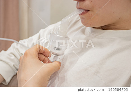 A close-up view of a person using a nebulizer, demonstrating the respiratory treatment process intended for asthma and other respiratory conditions. A close-up view of a person using a nebulizer, demonstrating the respiratory treatment process intended for asthma and other respiratory conditions. 140504703