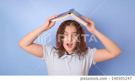 Playful Girl Balancing Book on Head Against Soft Blue Background, Expressing Joy and Curiosity Playful Girl Balancing Book on Head Against Soft Blue Background, Expressing Joy and Curiosity 140505247