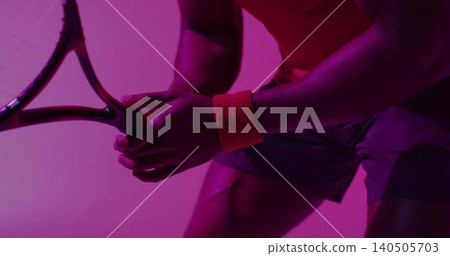 Gripping male athlete holding tennis racket in studio under magenta light, orange wristband, shorts Gripping male athlete holding tennis racket in studio under magenta light, orange wristband, shorts 140505703
