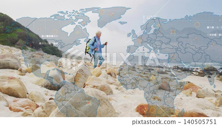Walking senior in blue jacket and leggings, using trekking pole on rocky shore with digital overlay 140505751