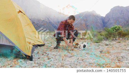 Kneeling camper in red plaid shirt reaching toward small terrier on pebbled plain with yellow tent Kneeling camper in red plaid shirt reaching toward small terrier on pebbled plain with yellow tent 140505755