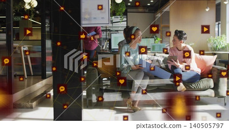 Laughing two women in light blouse, heels sharing tablet and documents in office lounge with icons Laughing two women in light blouse, heels sharing tablet and documents in office lounge with icons 140505797