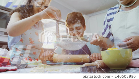 Sifting grandma with strainer while mom guiding boy rolling dough at kitchen counter, in aprons Sifting grandma with strainer while mom guiding boy rolling dough at kitchen counter, in aprons 140505986