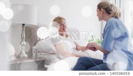 Smiling senior woman holding hands with caregiver in light-blue scrubs on bed at home with pillows Smiling senior woman holding hands with caregiver in light-blue scrubs on bed at home with pillows 140506058
