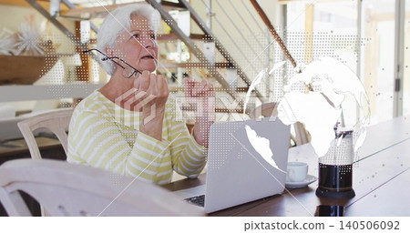 Talking senior woman holding glasses using laptop at home table with French press and globe overlay Talking senior woman holding glasses using laptop at home table with French press and globe overlay 140506092