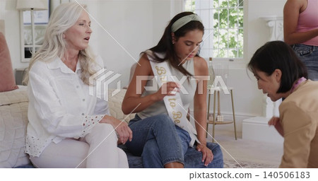 Adjusting seated woman wearing Bride to Be sash touching knee in living room, friends helping 140506183