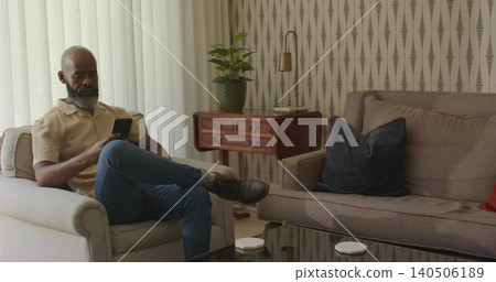Sitting middle-aged man holding smartphone in armchair in living room, wearing beige shirt 140506189
