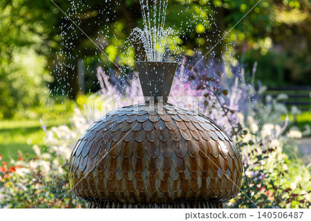 Water fountain releases droplets in a vibrant garden with blooming flowers on a sunny day. Water fountain releases droplets in a vibrant garden with blooming flowers on a sunny day. 140506487