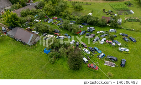 Numerous old cars are abandoned in a grassy area near several rustic buildings and gardens. Numerous old cars are abandoned in a grassy area near several rustic buildings and gardens. 140506488