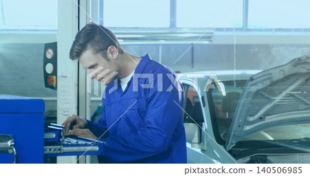 Inspecting mechanic in blue coverall, white tee leaning over tool cart at garage near silver car Inspecting mechanic in blue coverall, white tee leaning over tool cart at garage near silver car 140506985