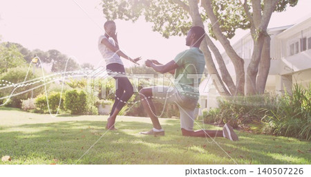 Kneeling man in green shirt offering object to woman in white top on lawn, dotted lights Kneeling man in green shirt offering object to woman in white top on lawn, dotted lights 140507226