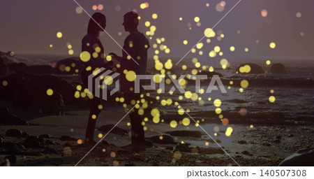 Holding hands backlit couple in dress and shirt facing on rocky shore with golden bokeh Holding hands backlit couple in dress and shirt facing on rocky shore with golden bokeh 140507308