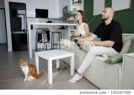 Playful young happy couple playing video games on sofa in modern white and green interior at home Playful young happy couple playing video games on sofa in modern white and green interior at home 140507406