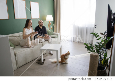 Indoor portrait of young happy couple playing video games on sofa in modern white and green interior Indoor portrait of young happy couple playing video games on sofa in modern white and green interior 140507408