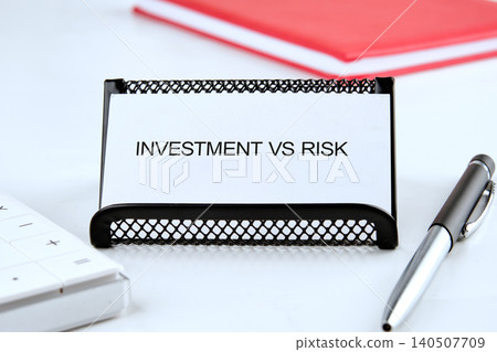 A card showing the words investment vs risk sits on a desk next to a calculator and a pen. A red notebook is visible in the background A card showing the words investment vs risk sits on a desk next to a calculator and a pen. A red notebook is visible in the background 140507709