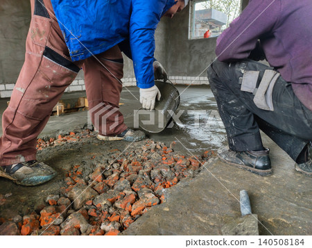 Workers pouring cement mix from a bucket onto a rough surface of broken bricks and concrete, creating a level foundation for a house renovation project. 140508184
