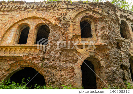 Crumbling walls of a brick building stand among trees. Openings show empty spaces inside. Nature surrounds the old structure Crumbling walls of a brick building stand among trees. Openings show empty spaces inside. Nature surrounds the old structure 140508212