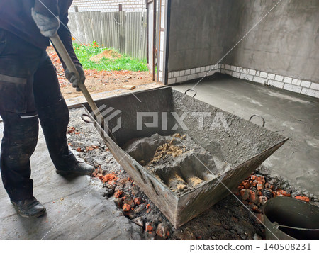 Construction worker preparing construction material, mixing cement mortar and sand with a shovel in a metal tank, a concrete floor curing in the background. Construction worker preparing construction material, mixing cement mortar and sand with a shovel in a metal tank, a concrete floor curing in the background. 140508231