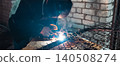 Welder wearing a protective helmet and gloves, joining metal parts of a chain-link fence, generating intense light and sparks during the welding process against a brick wall background. 140508274