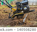 Person using a powerful walk-behind tractor to intensely cultivate freshly turned soil in a garden plot, preparing the ground for planting and seasonal agriculture. 140508280